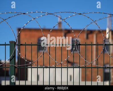 Grüne stahl Zaun mit Stacheldraht auf der Oberseite und roten Backsteingebäude im Hintergrund (unscharf) Stockfoto