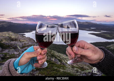 Cheers mit Rotwein über einem See in den Bergen mit einem rosafarbenen Sonnenuntergang. Natur, Reisen, Feiern, Konzepte. Stockfoto