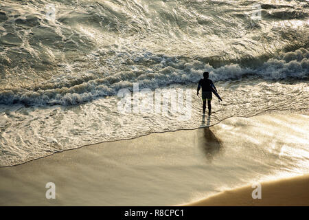 Ein Tourist mit seinem Surfbrett ist am Varkala Strand bei Sonnenuntergang bereit, das Meer zu gelangen. Indien. Stockfoto