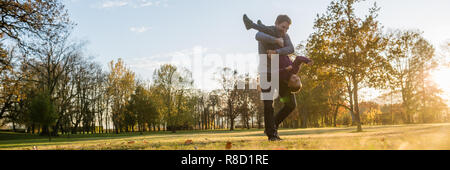 In der breiten Ansicht Bild einer jungen Vater Verklebung mit seinem Kleinkind Sohn in einem Herbst park ihn anheben und ihn mit der Oberseite nach unten drehen. Stockfoto