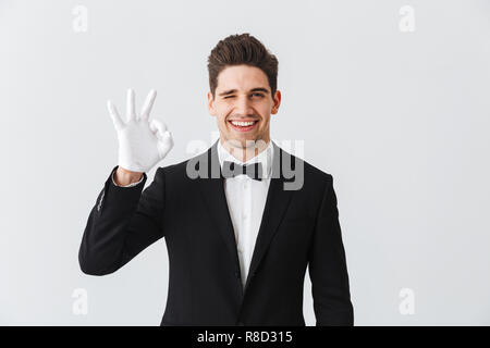 Porträt von einem gutaussehenden jungen Mann Kellner tragen Tuxedo und Handschuhe, die auf weißem Hintergrund, ok angezeigt Stockfoto