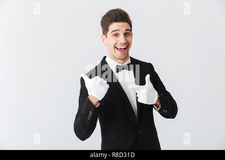 Porträt von einem gutaussehenden jungen Mann Kellner tragen Tuxedo und Handschuhe, die auf weißem Hintergrund, Daumen hoch Stockfoto