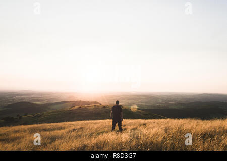 Ein einsamer Wanderer mit Blick über die englische Landschaft von einem Hügel auf einem Sommer Abend. Mit dem Sonnenuntergang, künstlerische Linsen Flare. Stockfoto