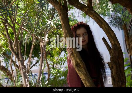 Ein hippie Mädchen mit Haar Blumen lehnt sich gegen einen Baum und sieht seitlich mit einem traurigen, besorgt und verwirrt Ausdruck Stockfoto