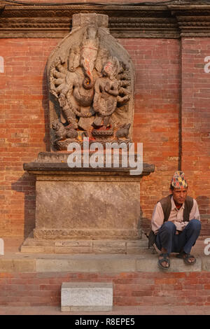 Ein Nepali Mann neben einem Relief mit der Darstellung eines multi-bewaffneten Version der hinduistischen Elefantengott Ganesh oder Ganpati in Durbar Suqare, Patan, Nepal sitzen Stockfoto