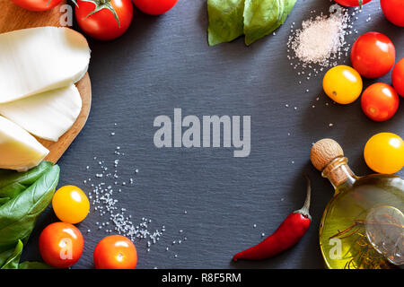 Tomaten, Mozzarella, Salz, Olivenöl und Basilikum liegen im Kreis auf dunklem Schiefer. Stockfoto