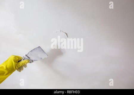 worker's hand in yellow gloves holding putty knife patching a hole with spatula with plaster or putty in white wall. Renovation and repair process, re Stockfoto