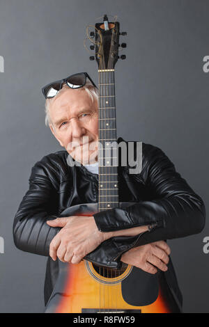 Älterer Mann in der Lederjacke und Sonnenbrille auf dem Kopf stehend isoliert auf Grau umarmen Gitarre suchen Kamera ernst Stockfoto