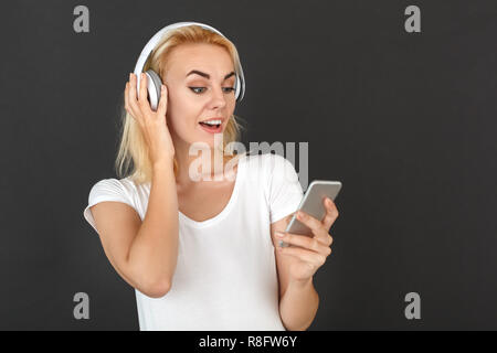 Freestyle. Frau in Kopfhörer steht auf schwarze Wand Auswahl Song auf dem Smartphone Musik isoliert überrascht Stockfoto