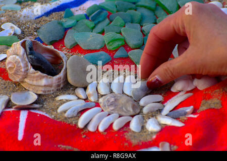 Muscheln - eine Vielzahl von Muscheln vom Strand. Muscheln, Meer farbigen Steine auf einem roten Handtuch auf den Sand. Sommer Strand Hintergrund. Blick von oben. Stockfoto