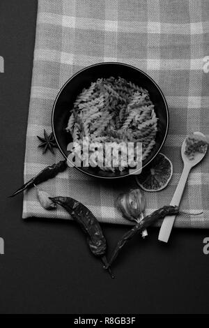 Leckere bunte Italienische Pasta in Keramik Teller mit Löffel aus Holz, Chili, Orange, Knoblauch, Sternanis und Zimt weiß und blau karierten Stoff oder karierten Tuch auf dunklem Hintergrund, Ansicht von oben Stockfoto