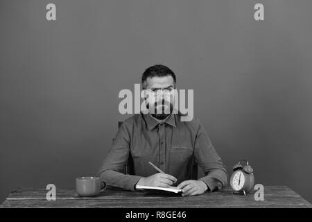 Schale, retro Wecker und rote Buch auf vintage Tabelle. Mann mit Bart und Brille schreibt im Notebook auf hellen roten Hintergrund. Prüfung und Studium Konzept. Mann mit ernsten Gesicht sitzt am Tisch Stockfoto