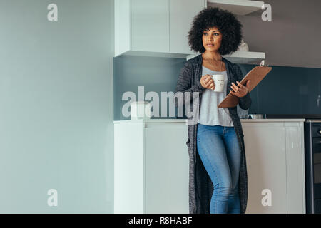 Junge Frau, die in ihrem Haus Küche mit Zwischenablage und Tasse Kaffee. Designer denken über neue Designs während der Arbeit im Home Office. Stockfoto