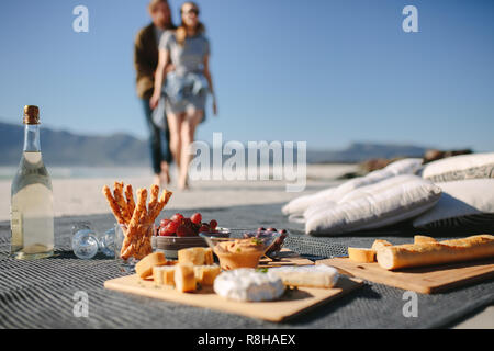 Essen und Trinken auf die Decke am Strand mit ein Paar auf dem Weg zu Ihm. Picknick am Strand mit Mann und Frau im Hintergrund. Stockfoto