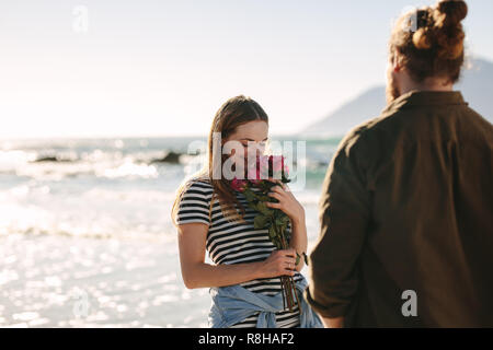 Attraktive Frau mit Blumen von Ihrem Freund mit dem Meer im Hintergrund. Zärtlich Paar auf romantisches Date am Strand. Stockfoto