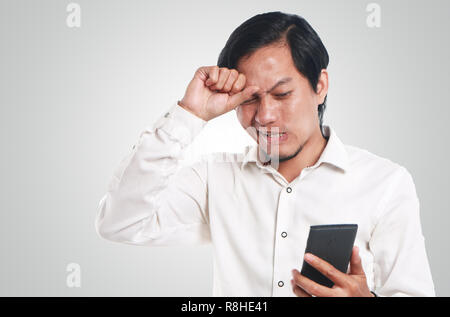 Foto Portrait eines lustigen jungen asiatischen Mann entsetzt, während Sie schlechte Nachrichten auf seinem Smartphone. Schlagen Kopf Halten Sie mit einer Hand das Telefon mit anderen h Stockfoto