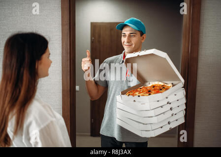 Fröhlichen Pizza Delivery boy zeigt Daumen nach oben Symbol für weibliche Kunden, schnelle Bereitstellung von Service. Kurier aus Pizzeria hält Karton Pakete im Innenbereich Stockfoto