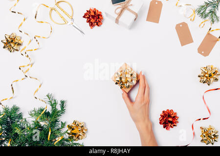 Die weibliche Hand, die eine kleine Geschenkbox mit goldenen Bogen unter flach Rahmen der grünen Tannenzweigen, Bänder und rote Weihnachtsschmuck. Stockfoto