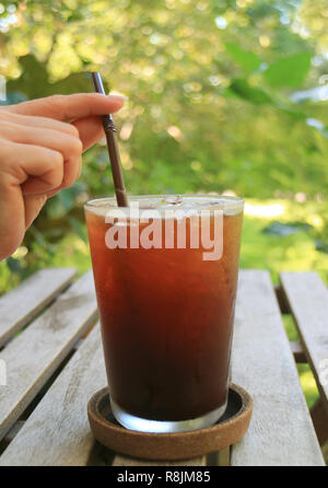 Weibliche Hand mit einem Strohhalm in Eiskaffee Glas mit unscharfen grünes Laub im Hintergrund Stockfoto