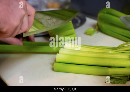 Der Küchenchef bereitet Selleriestangen mit einem großen Messer auf einem weißen Schneidbrett in einem Restaurant. Nähe zu sehen. Nur Hände. Stockfoto