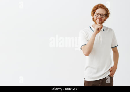 Charmant und verspielter Kerl gefragt sein Geheimnis sicher zu halten. Entspannt fröhlich und niedlichen Rothaarigen männliches Modell mit Bart Neigekopf lächelt freundlich sagen Shh, psst Geste mit dem Zeigefinger über Mund Stockfoto