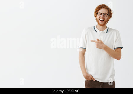 Studio shto des glücklichen und freudigen freundlichen redhead männliche Kursteilnehmer mit Bart in Gläsern und weiss Poloshirt die Hand in der Tasche zeigt mit dem Zeigefinger der linken und das Lachen aus Freude und gute Laune Stockfoto