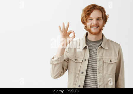 Hervorragende Arbeit, stolz auf unser Team. Portrait von zufriedenen gut aussehende freundlich und unbeschwert Rothaarige bärtige Mann in beige Jacke mit "ok" oder "Okay unterzeichnen und breit grinsend in perfekter Stimmung Stockfoto