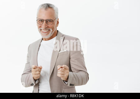 Het Gotcha. Portrait von amüsiert und glückliches sorgenfreies Alter Mann Gefühl erregt bereit, Geld in die Kamera zeigt mit Finger gun Gesten und lächelnd positiv in gute Stimmung über graue Wand zu machen Stockfoto