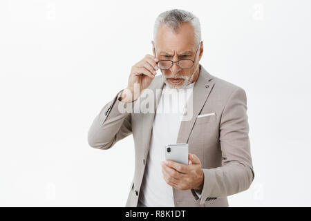 Portrait von verwirrten alten Mann, der versucht, zu verstehen, wie neue Smartphone, Handy, Brille konzentriert und stehen über grauer Hintergrund in der Klage in Frage bestimmt verwenden Stockfoto