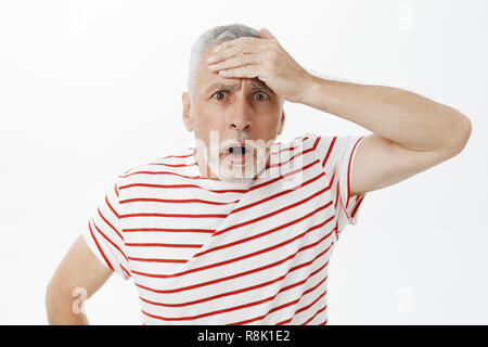 Portrait von geschockt, sprachlos, alte Mann stanzen Stirn mit Palm mit unruhigen Blick offenen Mund und Kamera unruhigen in schreckliches Problem über graue Wand gestört Posing Stockfoto