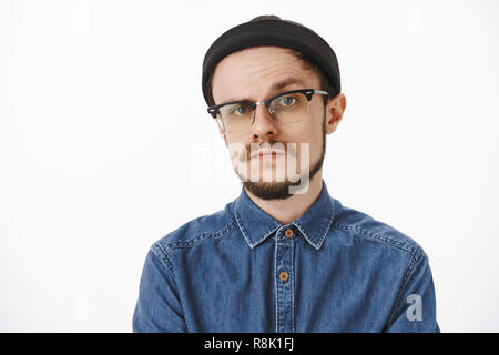 Zweifelhaft und ungewiss, Ernst - auf der Suche bossy männliche Unternehmer in Schwarz beanie Shirt und Gläser heben eine Augenbraue in Zweifel und Unglauben ständigen unemotive und Verdächtigen über grauer Hintergrund Stockfoto