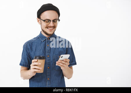 Taille-up Shot der geselligen glücklich und entspannt Junge stilvolle, moderne und Unrasierten Kerl in Gläser schwarz Beanie und blaues Hemd holding Smartphone und Papier Tasse Kaffee auf das Gerät mit dem Bildschirm lächelte freudig Stockfoto