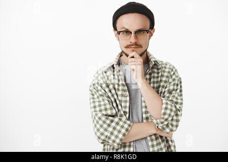 Taille-up Shot von interessierten kreativen bärtiger junger Mann in Schwarz Mütze und Brille die Hand am Kinn und starrte mit ernsten nachdenklichen Ausdruck an Kamera denken hören neugierig Plan Stockfoto