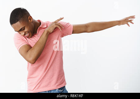 Glücklich und charismatische freudige afrikanischen amerikanischen Jungen Kerl, dab ziehen, Hände nach rechts kippen zufrieden, mit breiten glückliches Lächeln tanzen, Spaß haben über grauer Hintergrund Stockfoto