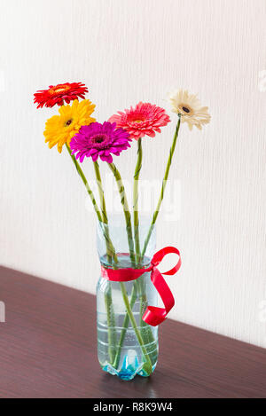 Fünf bunten Gerbera Blumen in einem plasctic Trinkflasche mit einem roten Band, auf einem Holztisch Stockfoto
