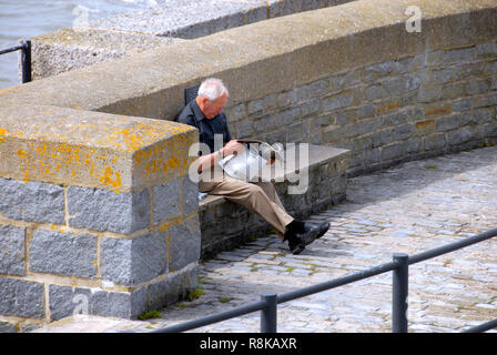 Mann sitzt auf Stein Sitz mit dem Rücken zum Meer an der Wand lesen Zeitung/Zeitschrift, Lyme Regis, Dorset, England Stockfoto