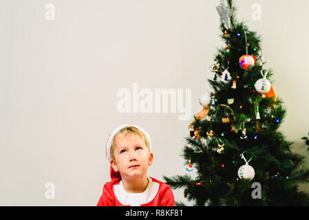 Blond und hübsch 5 Jahre alter Junge verkleidet als Weihnachtsmann durch den Weihnachtsbaum Langeweile beim Warten auf die Ankunft der Real Santa, weißer Hintergrund Stockfoto