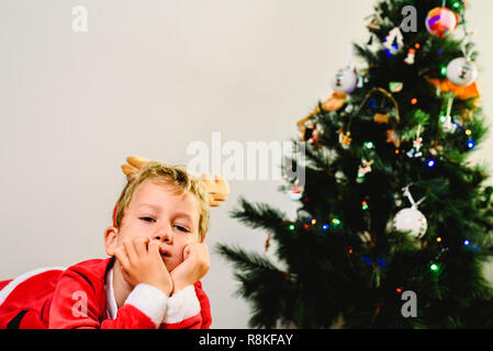 Blond und hübsch 5 Jahre alter Junge verkleidet als Weihnachtsmann durch den Weihnachtsbaum Langeweile beim Warten auf die Ankunft der Real Santa, weißer Hintergrund Stockfoto