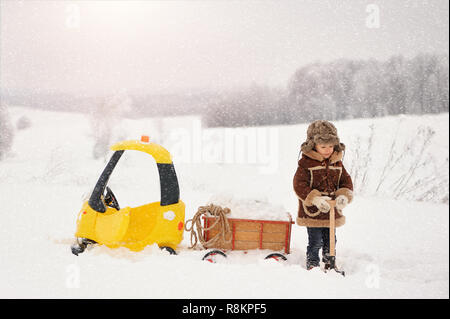 Happy Little Boy in Winterkleidung. Das Kind ist Spielen im Freien im Winter. Ein gelbes Spielzeugauto und eine kleine Warenkorb gefüllt mit Schnee, im Schnee stecken Stockfoto