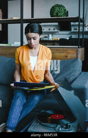 Attraktive gemischten Rennen Mädchen im orangefarbenen T-shirt Auswahl Vinyl auf dem Sofa im Wohnzimmer. Stockfoto