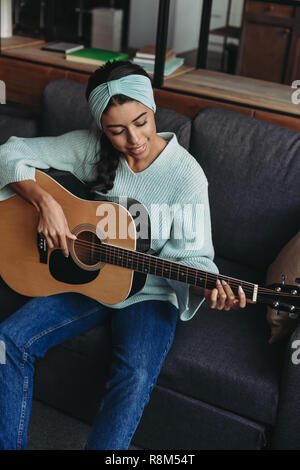 Hohe Betrachtungswinkel und der schönen gemischten Rennen Mädchen in türkis Pullover und Stirnband spielen akustische Gitarre auf dem Sofa zu Hause Stockfoto