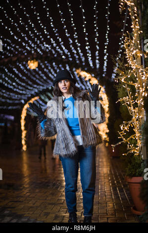 Hübsche dunkelhaarige Mädchen mit einem Pelzmantel, blaue Jeans, blau oben und einen schwarzen Hut, lächelnd, mit Schneeflocken Christmas lights Outdoor bei Nacht ti Posing Stockfoto