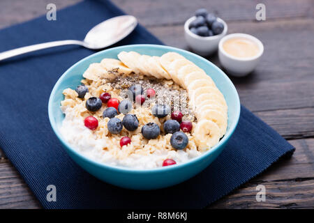 Gesunde fitness Frühstück: Haferflocken mit Bananen, Blaubeeren und Preiselbeeren, Chia Samen und Joghurt auf dunklen Holzplatte mit Textur Stockfoto
