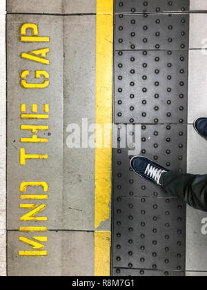 Füße treten auf ruber Oberfläche hinter gelben Safety Line der Londoner U-Bahn. Mind the gap Zeichen auf dem Boden geschrieben Stockfoto