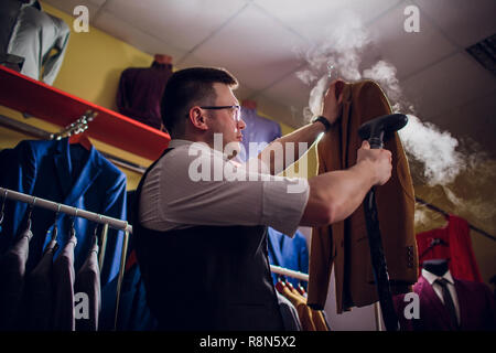 Individuelle Schnitte und näht einen Anzug. Ein junger Mann in der Schneiderei shop macht die Kleidung der Männer. Maßgeschneiderte Bekleidung Werkstatt. hält in ihrer Hand ein elektrisches Bügeleisen. wet - Wärmebehandlung Stockfoto