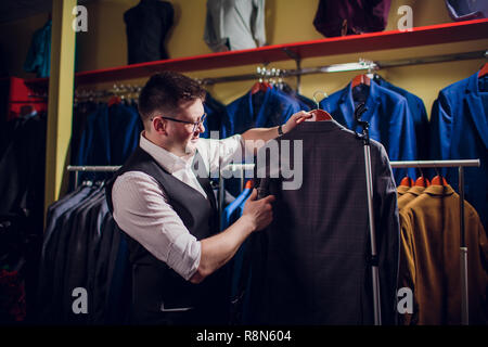 Individuelle Schnitte und näht einen Anzug. Ein junger Mann in der Schneiderei shop macht die Kleidung der Männer. Maßgeschneiderte Bekleidung Werkstatt. hält in ihrer Hand ein elektrisches Bügeleisen. wet - Wärmebehandlung Stockfoto