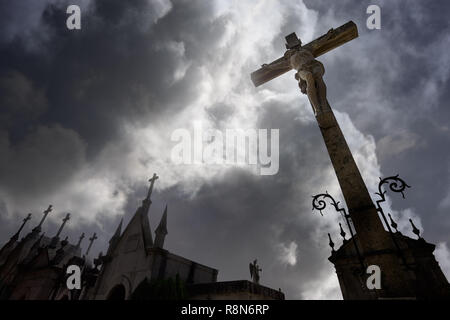 Alten, dunklen europäischen Friedhof gegen einen dramatischen bewölkten Himmel mit einem gekreuzigten Christus Statue auf der Vorder- und mehrere Gräber in Form von Kapellen. So Stockfoto