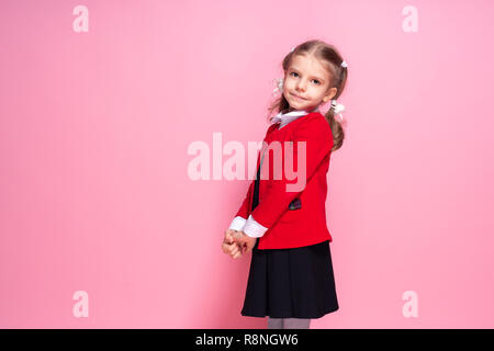 Adorable kleine Mädchen in der Roten Schule Jacke und schwarzes Kleid mit Blick auf die Kamera beim Stehen auf rosa Hintergrund Stockfoto