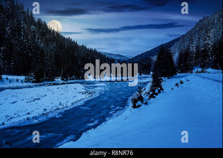 Mountain River im Winter in der Nacht bei Vollmond Licht. Schnee bedeckten Ufer. Wald im Schnee auf die fernen Berge. bewölkt Morgen Stockfoto