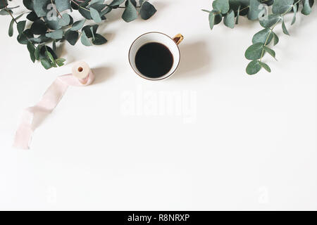 Blumige Komposition aus grünem Eukalyptus Blätter und Zweige auf weißem Holz- Hintergrund mit Tasse Kaffee und Seidenband. Feminine Büro Schreibtisch, eingerichteten Lager Bild. Flach, Ansicht von oben. Stockfoto